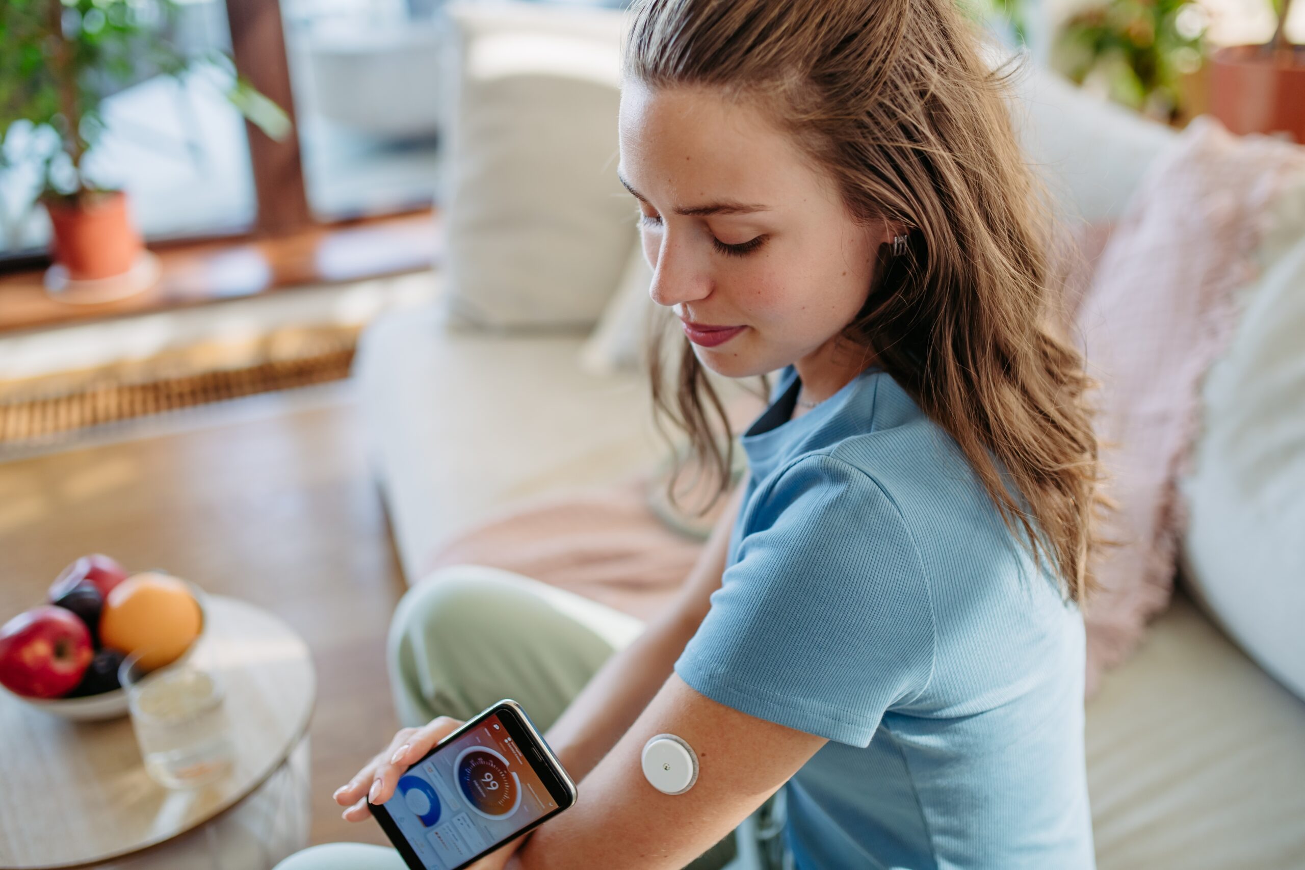 <p>The image depicts a young woman with long, light brown hair casually seated on a light olive green cushion on a wooden floor in a brightly lit, comfortable living room. She is focused on her smartphone, which she holds in her left hand, and appears to be scanning it with a device positioned over her upper left arm. The device, a small circular patch, is likely a continuous glucose monitor (CGM). The phone screen displays a circular graph with a number ’99’ prominently visible, suggesting a blood glucose reading. She is wearing a light blue t-shirt and her expression is one of calm concentration. The background features a warm, inviting atmosphere with a large window allowing natural light to flood the room. A woven rug is partially visible, and a potted plant sits on a windowsill. A small round table beside her holds a bowl of fruit – apples and peaches – and a decorative plate. A plush, textured pillow in shades of pink and cream is visible behind her, adding to the cozy ambiance. The overall impression is one of health management integrated seamlessly into a relaxed, everyday lifestyle, showcasing technology aiding in personal wellness within a comfortable home setting.</p>” style=”display: block; margin-left: auto; margin-right: auto; border-radius: 5px; max-width: 80%; height: auto;” loading=”lazy”/><figcaption>(<em>img by Institute of Nutrition and Fitness Sciences – INFS</em>)</figcaption></figure><p>This can require adjustments in diabetes medications or insulin dosing and affect <a href=